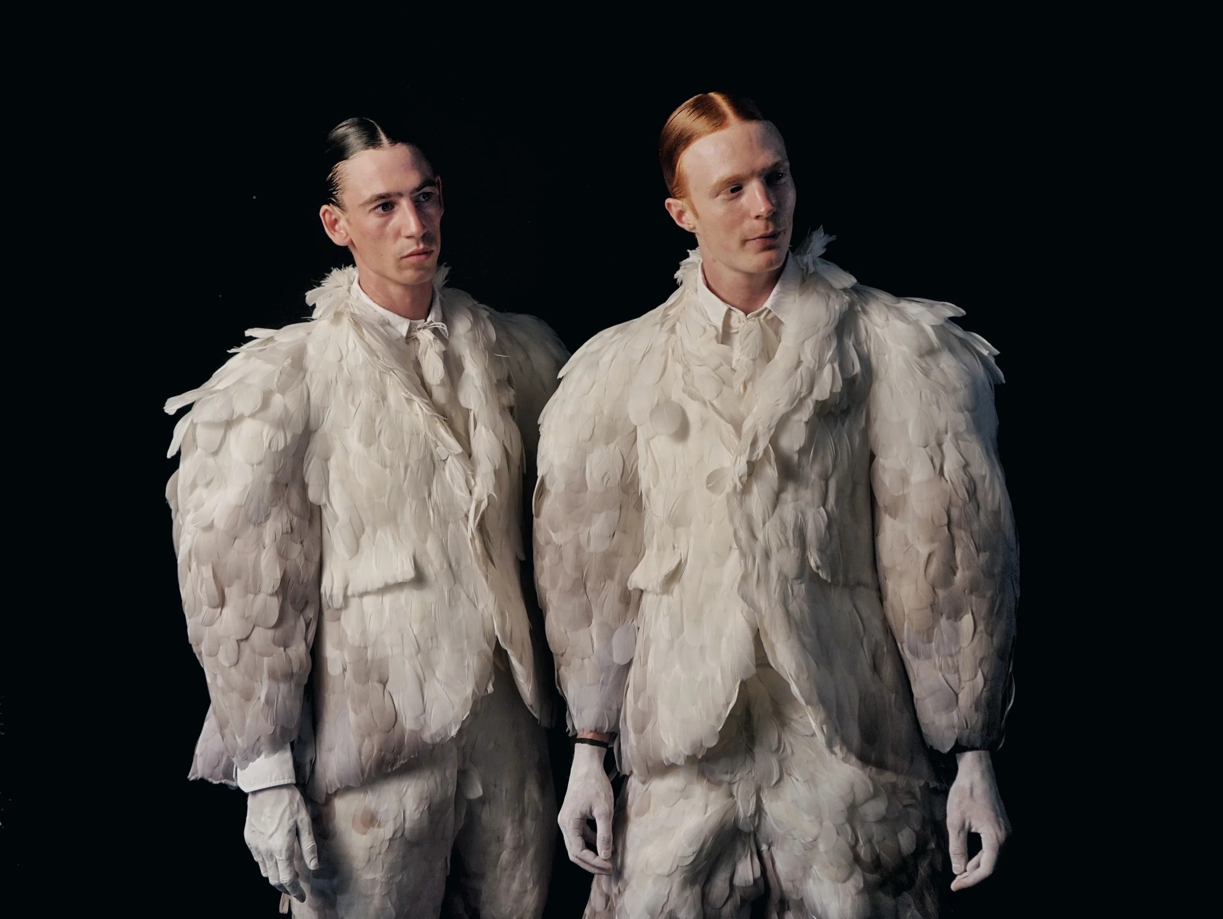 Two models wearing white feathered outfits against a dark background
